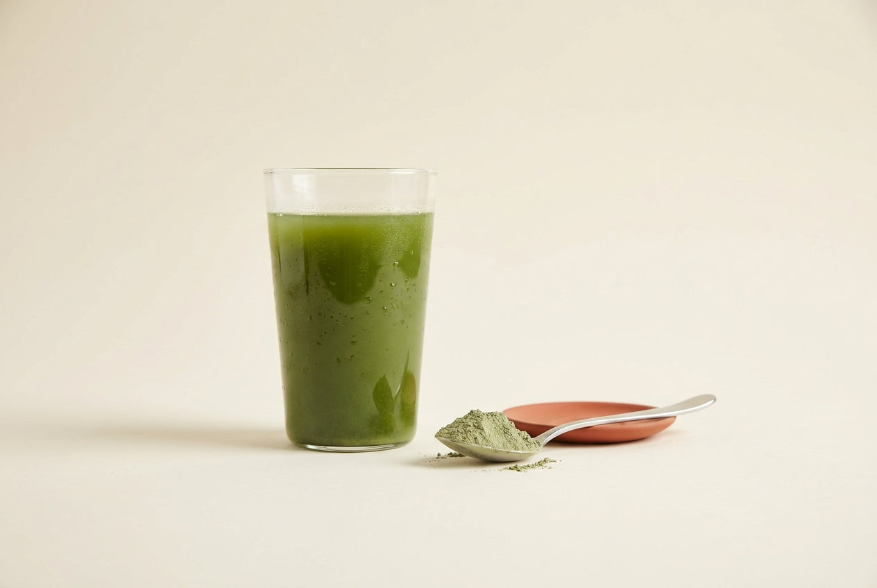 Tall glass of green drink beside a spoon of green powder on a cream background